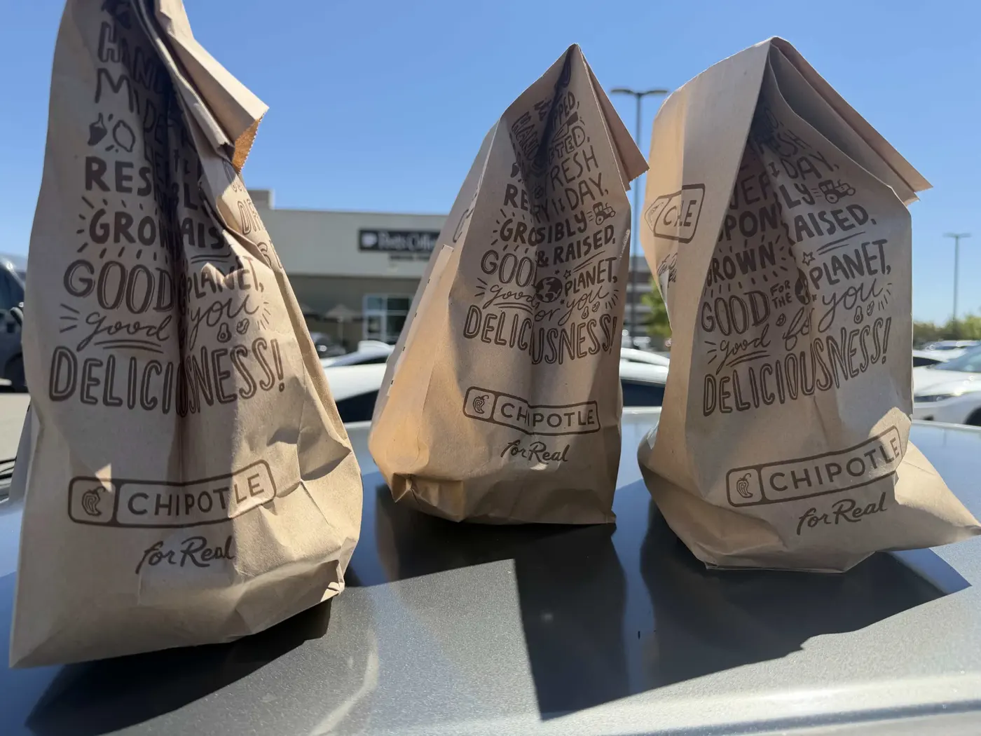 Three Chipotle takeout bags lined up on a car roof in the sun
